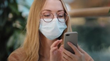 Gözlüklü beyaz kız yüz koruma maskesi takıyor. Cep telefonlu kadın ekranına bakıyor. Beklenmedik haberlerin şokunu yaşıyor.