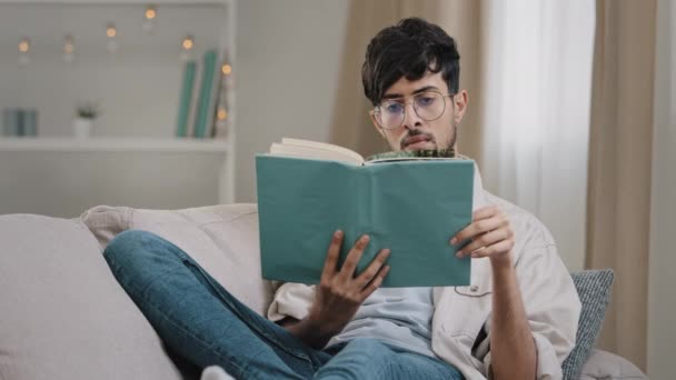 Jovem estudante inteligente concentrado árabe hispânico homem árabe masculino usa óculos deitado no sofá em casa leitura livro apreciando estudo de literatura com livro didático em casa, conceito de aprendizagem hobby — Vídeo de Stock