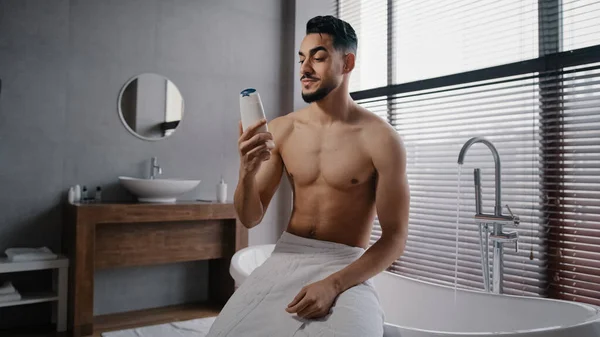 Árabe Indiano cara muscular nu sexy homem sem barba usa toalha de banho branca nos quadris senta-se no banheiro se preparando para tomar banho de lavagem farejando gel de banho refresco matinal com cosméticos masculinos naturais — Fotografia de Stock