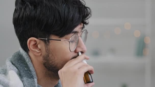 Close-up arabian indian bearded guy with glasses lonely sad sick ill man wrapped in blanket spraying rhinitis spray anti-allergic remedy drug in nose cold medicine seasonal allergy flue symptoms — Video
