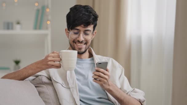 Молодой расслабленный парень индийский арабик бородатый красивый мужчина сидит дома на диване пьет чай кофе из чашки глядя на телефон улыбаясь смотреть смешное видео в чате с помощью смартфона — стоковое видео