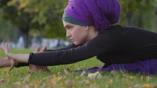 Femme musulmane en hijab se trouve sur la pelouse d'herbe verte dans les virages du parc dans le corps faisant plier femme étirement yoga entraînement sport pratique exercice méditation équilibre effort étirer les jambes. Fille flexion flexibilité — Video