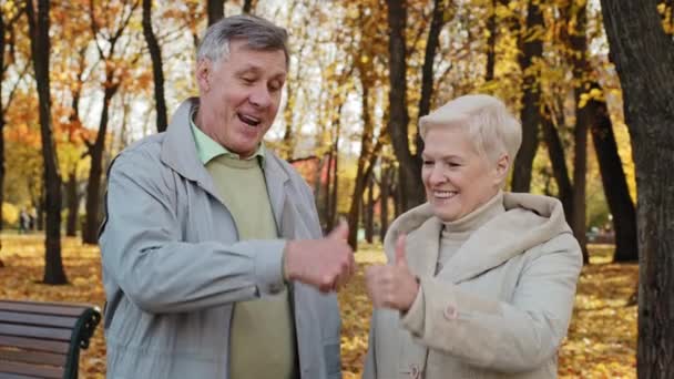 Porträtt av glad äldre kaukasiska familj hustru make äldre morföräldrar står utomhus i höst park leende gamla mogna par senior man kvinna visa tummen upp hand gest godkännande rekommendation — Stockvideo