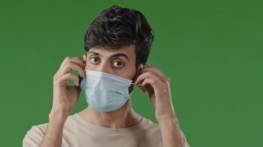Milenyum bitkin esmer erkek hasta Arap öğrenci yeşil arka planda duruyor. Tıbbi maske takıyor, nefes alıyor, derin nefes alıyor salgının son nefesini kesiyor.