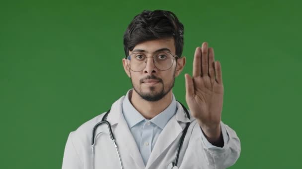 Ung manlig läkare man i medicinsk mask isolerad i grön chroma nyckel studio visa stopp tecken med oöverkomlig hand gest säkert avstånd sätter handflatan framåt oense stoppa coronavirus infektion sprider — Stockvideo