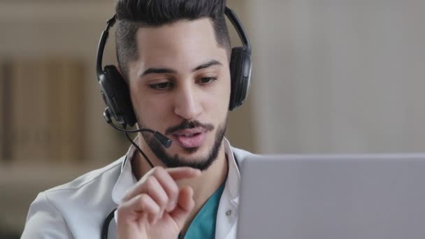 Professionele mannelijke Arabisch Spaanse jonge arts assistent in medische jas dragen headset met microfoon te maken conference call op laptop gebruik afstand virtuele leren computer app online te raadplegen in video chat — Stockvideo