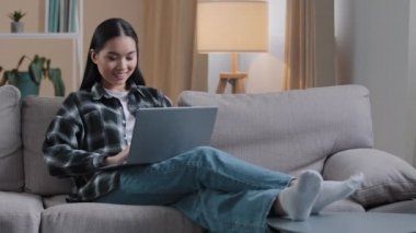 Gülümseyen Asyalı kadın bilgisayarını kullanarak internette bilgisayar oyunu arıyor. Koreli kız öğrenci kanepeye oturmuş e-öğreniyor internet eğitimi alıyor Wi-Fi ile çalışıyor.