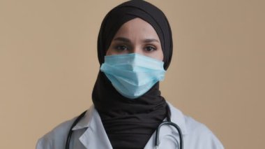 Arap Müslüman kadın hemşire portresi tesettürlü koruyucu bir maske takıyor. Steteskopla hastanede poz veriyor. Klinikteki genç kadın doktor kameraya bakıyor.