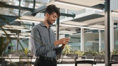 Gülümseyen mutlu bin yıllık Arap erkek profesyonel İspanyol işadamı akıllı telefon parşömeni cep telefonlu cep telefonlu cep telefonu mesajını ofiste tutuyor sosyal uygulamalarda mobil sohbet kullanarak interneti düzene sokuyor.