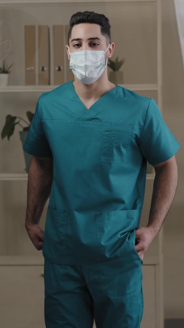 Вертикальный вид араб латиноамериканец молодой парень в защитной медицинской маске врач медсестра хирург в униформе глядя на позирование камеры положил руки в карманы стоять в больнице во время эпидемии ковид-19 — стоковое видео