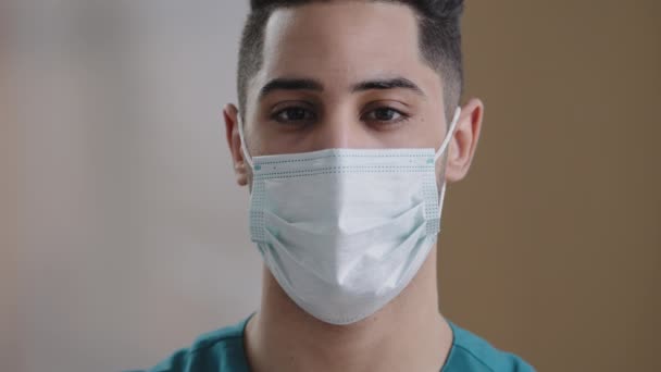 Primer plano joven hispano doctor árabe cara masculina en máscara médica con ojos oscuros mirando cámara confiado adulto chico profesional enfermera trabajo en oftalmología cirugía vista corrección operación — Vídeos de Stock
