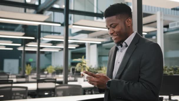 Glücklich millennial mann afrikanisch-amerikanisch männlich unternehmer mitarbeiter erwachsener kerl halten handychat in sozialen netzwerken surfen internet verwenden handysuche informationen hochladen online lustige video im büro — Stockvideo