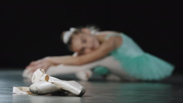 焦点を当てないぼやけた少女アーティストダンサー疲れた子供のバレリーナダンスの後に床に座って足の筋肉を伸ばし柔軟性をストレッチ疲れポイントシューズバレエの靴を見て踊って後 — ストック動画
