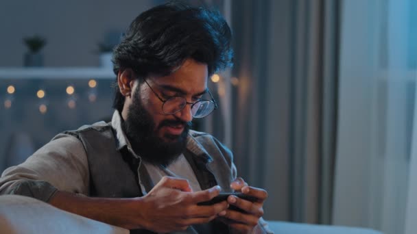 Millenniumi 30-as férfi szakállas szemüveg ül otthon késő este rabja online játékok mobil szórakoztató alkalmazás olvassa hírek böngészés telefonos srác gamer okostelefon játék app — Stock videók