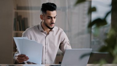 Ciddi Arap genç iş adamı erkek profesyonel çalışan bilgisayar klavyesi fotokopisi kağıt metin elektronik form odaklı Hispanik adam belge karşılaştırma online mali rapor