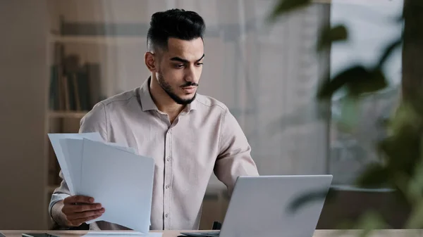 Serious arabian young business guy male professional worker employee type on computer keyboard copy paper text at electronic form focused hispanic man compare documents check online financial report — стоковое фото