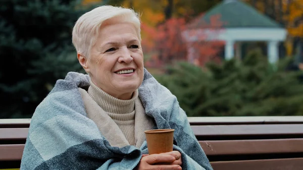 Feliz maduro única mulher de meia idade coberto com xadrez quente beber café quente copo descartável saudável alegre aposentado sênior desfrutando de descanso ao ar livre sorrindo velha senhora sentada no banco do parque de outono — Fotografia de Stock