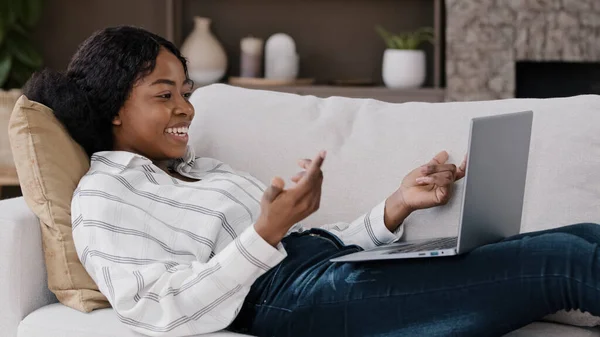 Evdeki kanepede uzanan Afrikalı kadın laptoptan selamlama ekranına bak. Yurtdışında yaşayan aile dostumuzla uzaktan konuşma görüntüsü al. Amerikalı kadın konferansla iletişim kuruyor. — Stok fotoğraf