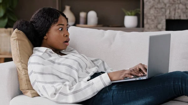 Menina africana estudante usuário freelancer encontra-se no sofá em casa olhando para laptop usando internet verifica e-mail lê má notícia inesperada sente surpresa choque olhando para câmera abre a boca profundamente de espanto — Fotografia de Stock