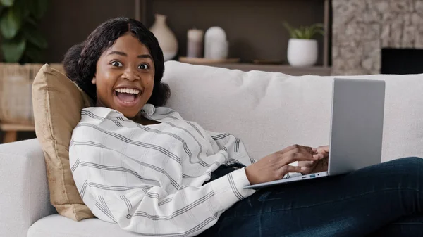 Kanepeye dizüstü bilgisayarı olan Afrikalı kız öğrenci iyi haberler alıyor. Test işi için yüksek not teklif ediyor. Mutluluk başarısı. — Stok fotoğraf