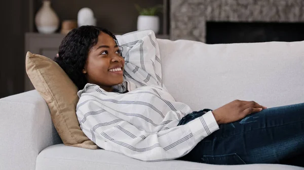 Mutlu, rahat, Afro-Amerikan kadın dinlenme odası kanepede uzanıp huzur dolu bir hafta sonu dinlenme keyfine varıyor. Oturma odasında dinleniyor. Tembel, sakin bir kız, evinde rahat bir mobilyada gülümsüyor. — Stok fotoğraf