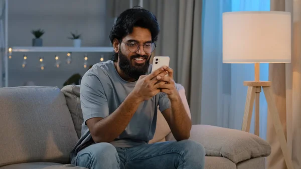 Χαμογελώντας Arabian ινδική άνθρωπος γενειοφόρος αρσενικό ψάχνει στο χαμόγελο smartphone γραπτών μηνυμάτων με φίλη μηνυμάτων κουβέντα στο σπίτι κάθεται στον καναπέ το βράδυ παίζοντας το παιχνίδι στο κινητό app βλέποντας αστείο βίντεο — Φωτογραφία Αρχείου