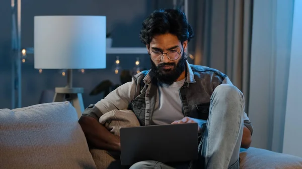 有识之士，阿拉伯裔男性自由职业者，印地安人，留着胡子的成年人，思考着计算机项目问题的解决方案，在笔记本电脑上寻找答案，在家中寻找答案，怀疑男性在深夜远程工作 — 图库照片