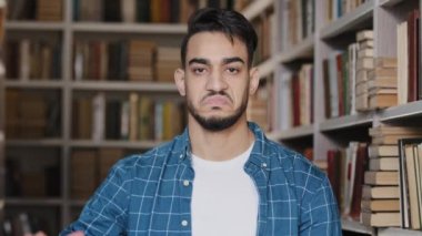 Yakın plan hoşnutsuz Hintli öğrenci kütüphanede kameraya bakıyor. Onaylamadığını gösteriyor. Eğitimden memnun olmayan düşük puanlı jest başarısızlığı.