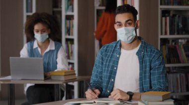 Kütüphanede oturan öğrenciler maske takmış genç mutlu adamı çalışırken masaya oturup ödev hazırlıyorlar gülümseyen kameraya bakıp selamlar sallıyorlar yüksekokul konseptini selamlıyorlar