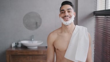 Banyodaki komik erkek portresi İspanyol Hindistanlı çıplak adam sakallı adam beyaz sabunlu köpükle gülümsüyor sakal traş jeli tıraş jeli ile banyo havlusu tutuyor kameraya bakıyor traş olmaya hazırlanıyor