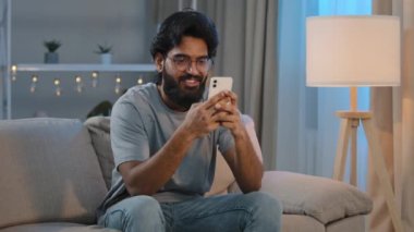 Gülümseyen Arap Hintli sakallı adam akıllı telefonlu gülüşüne bakıyor kız arkadaşıyla mesajlaşıyor, sohbet ediyor geceleri kanepede oturuyor mobil uygulamalarda oyun oynuyor ve komik video izliyor.