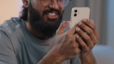 Kesik manzaralı, tanınmayan, sakallı, akıllı bir adam elinde telefonla gülüyor, dişlek gülüyor, mesajlaşıyor, evde komik videolar izliyor, internette sohbet ediyor ve mobil bir sosyal medya gülüşü kullanıyor.