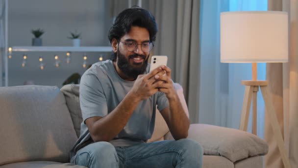 笑顔アラビア語インディアン男Beaded男性見ますでスマートフォンの笑顔テキストメッセージで友人ガールフレンドメッセージングチャットで自宅で座っていますソファに夜遊びゲームでモバイルアプリを見て面白いビデオ — ストック動画