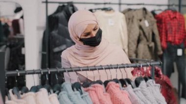 Medikal koruyucu maskeli genç Arap kadın alışverişçi elbise mağazasından indirimli satış indirimi alışverişi için butik müslüman kadın müşteri tesettürlü yeni sıcak süveter arıyor