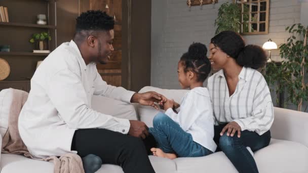 アフリカ系アメリカ人の親は子供の娘とソファに家に座って時間を過ごす リビングルームでの出産家族はソファでリラックスした笑いの結合関係カジュアルな会話を話しています — ストック動画