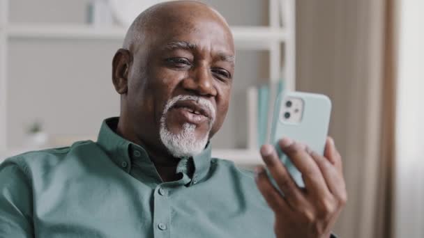 オフィスソファに座っている高齢者60年代のビジネスマンは モバイルアプリとビデオ通話を持っています 古いですアフリカ系アメリカ人男性保持スマートフォンを呼び出しますオンライン会議チャットリラックス上のソファ ホーム — ストック動画