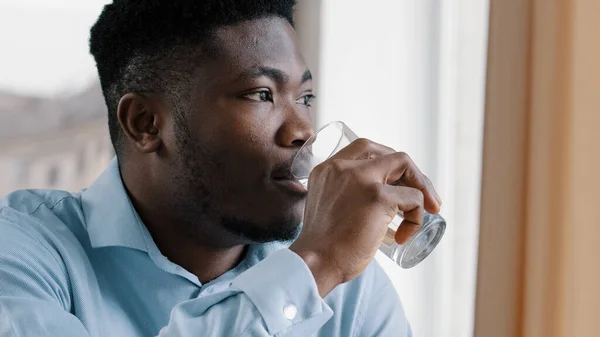 African american young dreaming man thoughtful positive guy turn head look at window drink fresh cold water glass of pure liquid hydrate thirst suffer from hot make pause feeling rejuvenation energy