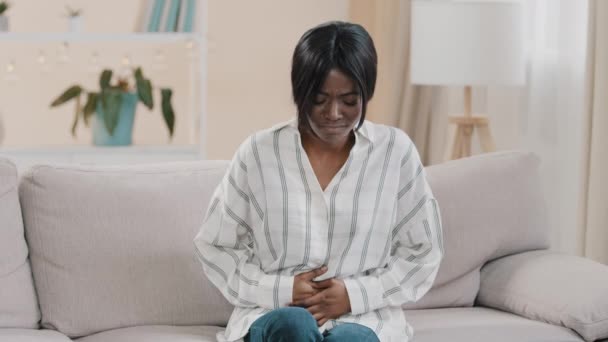 若い動揺不健康なアフリカ系アメリカ人女性に座っているソファの上に座っている胃の上に手を握って腹部の痛み感からの痙攣月経痛感症状吐き気 — ストック動画