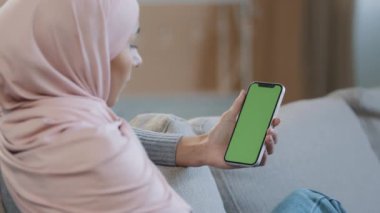 Omuz üzerinden İslami Arap kadın tesettürlü rahat koltuğa oturup yeşil ekran kroma anahtar ekranı ile sosyal uygulama hizmetleri web sitesini kullanarak el ele tutuşuyor.