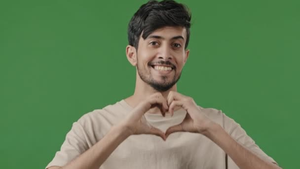 笑着他的恐慌Arab快乐的男人印地安人男友看着摄像机显示慈善爱心手势手指头心形浪漫的标志仁爱的象征站在绿色工作室的彩色按键上 — 图库视频影像