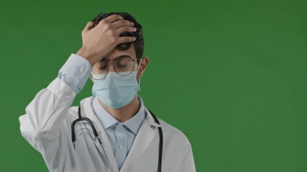 惊慌失措的形象年轻沮丧的印度男人用手捂住额头疲倦的医生蒙面失败愤怒的坏消息神经质站在绿色的背景上 — 图库视频影像