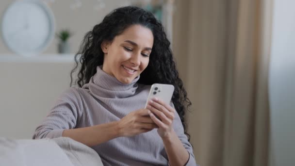 若い女性魅力的なアラビア語のイスラム教徒の女の子の女性でヒジャーブはソファの上に座って笑顔イスラム教徒の女性のテキストメッセージスマートフォンで彼氏と仕事をしますモバイルデジタルアプリショッピングオンライン検索情報 — ストック動画