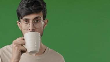 Arap Hindistanlı yakışıklı genç adamın yakın portresi yeşil arka planda duruyor sıcak bitki çayı içiyor soğuk su içiyor İspanyol erkek gündüz uykusunda dinleniyor.
