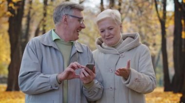 Sonbahar parkında yürüyen neşeli Kafkasyalı yaşlı çift akıllı telefon kullan mobil internet tarayıcı oku iyi haber izle komik video kaydırma sayfaları sosyal uygulamalarda yaşlı arkadaşlarıyla sohbet et