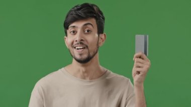 Tişörtlü mutlu Arap erkek bankacı çalışanın stüdyo portresi, plastik bankacılık kredi kartının internet üzerinden alışveriş yaptığını gösteriyor.