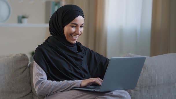 笑顔Arabイスラム教徒女性イスラム教徒若い女の子でHihabフリーランスの学生を使用してノートパソコンの傾きオンソファにホームタイプの電子メール仕事オンラインブラウジングネットゲームチャット友人自己教育 — ストック動画