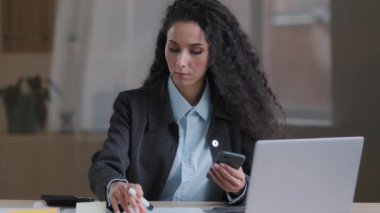 Odaklanmış Arap İspanyol genç iş kadını çoklu görev uzmanı kadın denetçi yapışkan notlar üzerine görevler yazar pazarlama planını düzenler cep telefonu arama bilgilerini kullan
