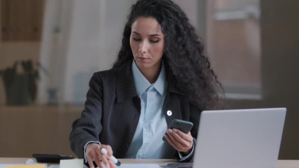 Fókuszált arab spanyol hispán fiatal üzletasszony multitasking specialista női könyvvizsgáló írni feladatokat ragacsos jegyzetek kezelése papírmunka szervezése marketing terv használata mobil eszköz mobiltelefon keresési információk