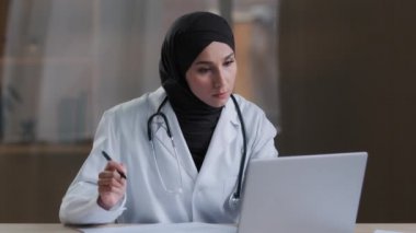 Yoğun Müslüman kadın doktor doktor doktor. Belgeleri imzalıyor. Klinik kabinesinde laptopta arama bilgileri bulunuyor.