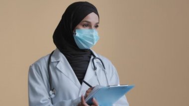Kadın Arap tıp çalışanı doktor, Müslüman terapist, hastane kliniğinde güvenlik maskesi başörtüsü takıyor, not tutuyor, sağlık sorunları, sağlık raporlarını dolduruyor, sigorta formunu dolduruyor.